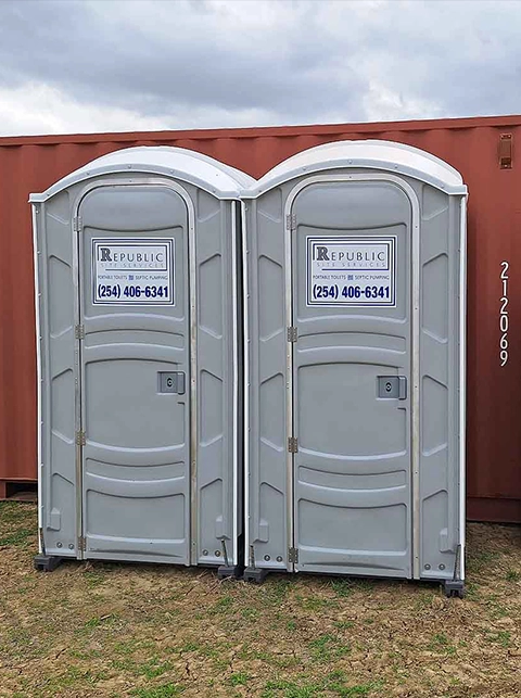 Two Gray Portable Toilet Rentals Outdoors on Grass Next to Red Container